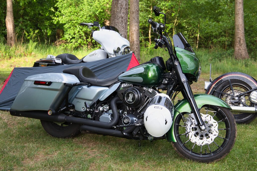 Ein glänzendes grün-silbernes Tourenmotorrad steht auf einer Wiese in der Nähe eines kleinen grauen Zelts, mit einem weiteren weißen Motorrad und Bäumen im Hintergrund.
