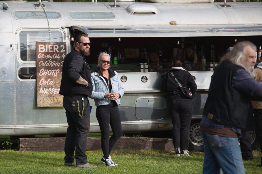 Ein Mann und eine Frau stehen lächelnd vor einem alten silbernen Imbisswagen, auf dem eine Getränkekarte ausliegt. Andere Menschen sind in der Nähe versammelt, und die Szene spielt sich im Freien auf einer Wiese ab.