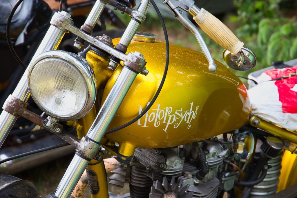Nahaufnahme eines alten gelben Motorrads mit der Aufschrift "Motorpsycho" in stilisierten Buchstaben auf dem Kraftstofftank. Der Scheinwerfer, der Lenker und ein Teil des Motors sind zu sehen.