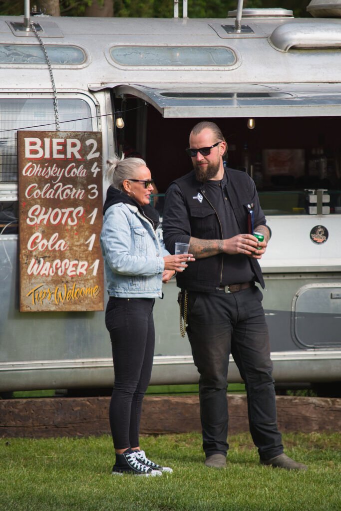 Zwei Personen in Freizeitkleidung stehen vor einem silbernen Imbisswagen und unterhalten sich. Auf einem Holzschild sind die Getränkepreise in deutscher Sprache angegeben. Beide halten Getränke in der Hand und tragen Sonnenbrillen. Die Atmosphäre wirkt entspannt und ungezwungen.