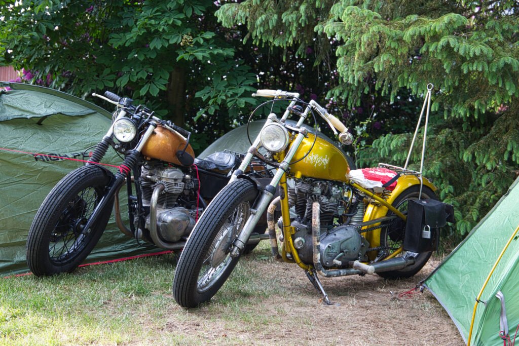 Zwei Oldtimer-Motorräder, ein braunes und ein gelbes, stehen im Gras zwischen grünen Campingzelten und Kiefern. Die Motorräder haben ein klassisches, individuelles Aussehen mit freiliegenden Metallteilen und ledernen Satteltaschen.