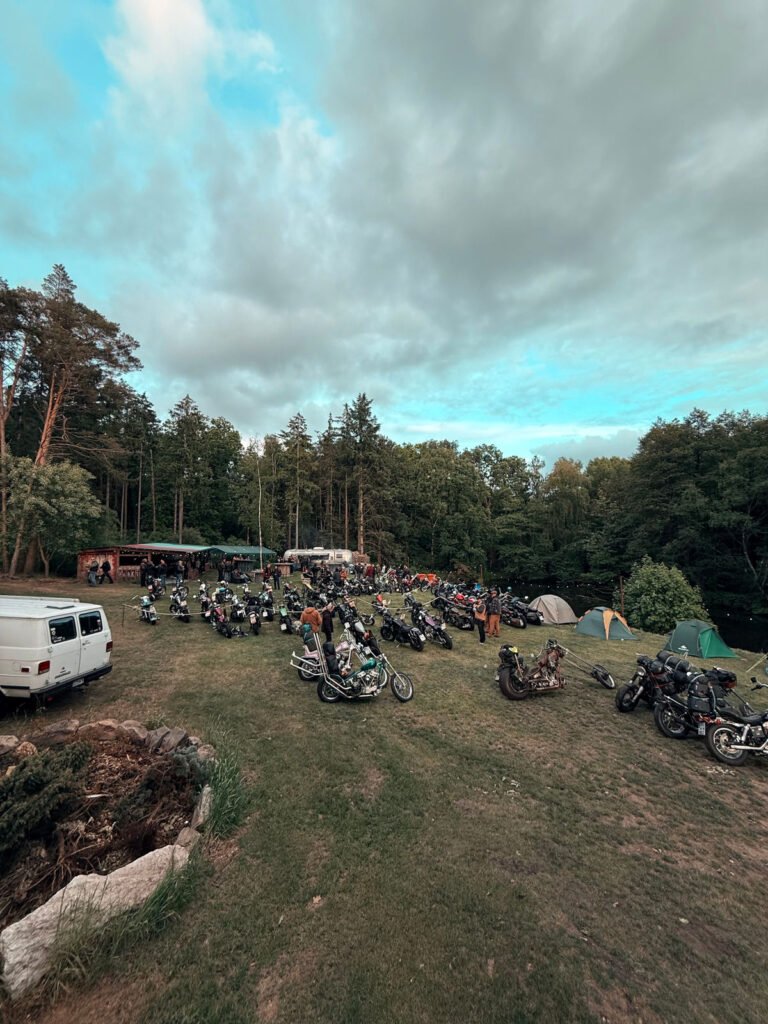 Eine grasbewachsene Lichtung mit zahlreichen geparkten Motorrädern, mehreren Zelten und einem weißen Lieferwagen, umgeben von Bäumen unter einem wolkenverhangenen Himmel, bei dem es sich offenbar um eine Camping- oder Motorradveranstaltung handelt.