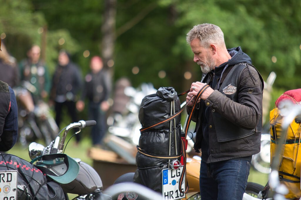 Ein Mann mit grauem Haar steht im Freien neben Motorrädern und sichert einen schwarzen Seesack mit Bungee-Seilen. Der Hintergrund ist grün und unscharf, was auf eine Versammlung oder Veranstaltung in einer parkähnlichen Umgebung hindeutet.