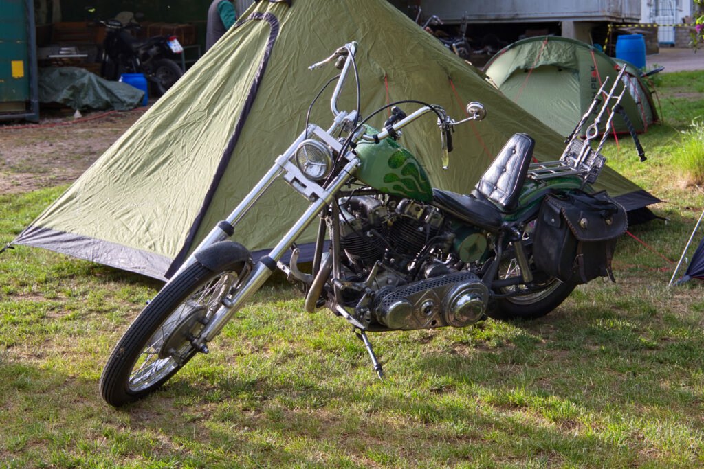 Ein Custom-Chopper-Motorrad mit hohem Lenker und grünem, geflammtem Tank ist auf einer Wiese vor einem grünen Campingzelt geparkt. Im Hintergrund sind Campingausrüstung und andere Zelte zu sehen.