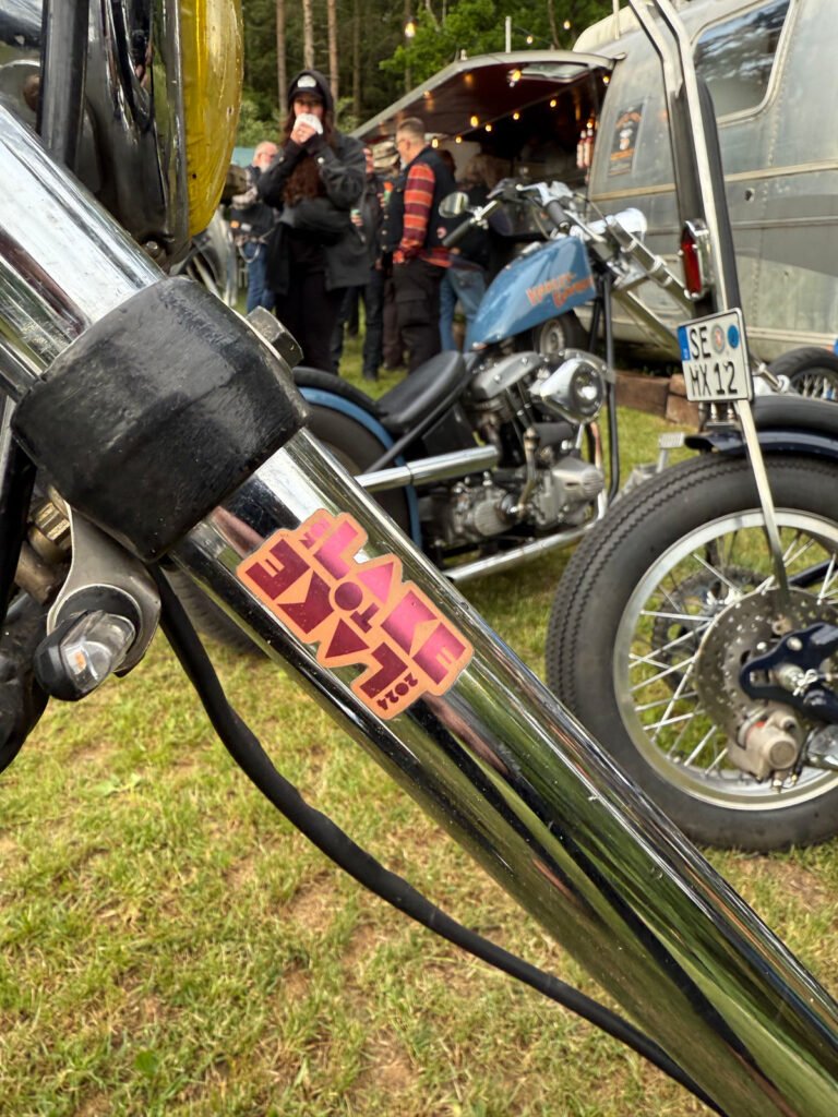 Nahaufnahme einer Motorradgabel mit einem "Have Fun"-Aufkleber, geparkt im Gras. Im Hintergrund stehen Menschen neben einem anderen Oldtimer-Motorrad und einem silbernen Anhänger bei einer Veranstaltung im Freien.