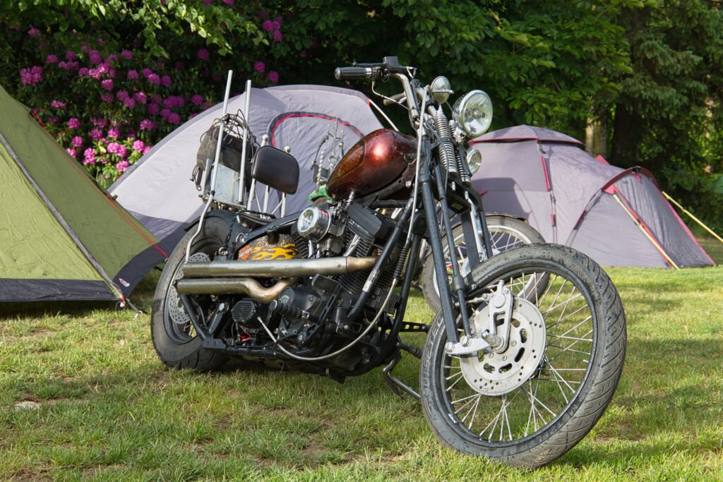 Ein Custom-Motorrad mit Flammendetails steht auf einer Wiese in der Nähe mehrerer Zelte, umgeben von Bäumen und blühenden Blumen.