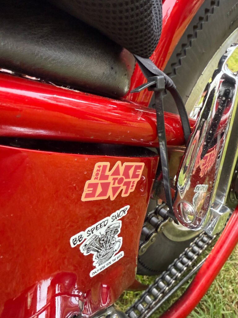 Nahaufnahme eines roten Motorradrahmens mit zwei Aufklebern. Auf dem einen steht in fetten Buchstaben "LAKE TO EMIT", auf dem anderen "BB SPEED SHOP" mit der Illustration einer geflügelten Zündkerze. Ein Reifen und ein Chromteil sind ebenfalls zu sehen.