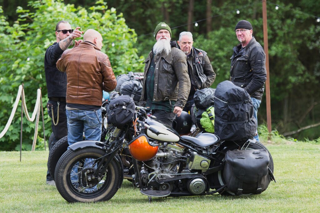 Fünf Männer in Lederjacken und Outdoor-Ausrüstung stehen neben zwei beladenen Motorrädern auf einer Wiese mit Bäumen im Hintergrund und scheinen auf einem Road Trip oder einer Versammlung zu sein.