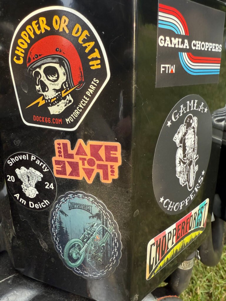 Eine schwarze Oberfläche, die mit bunten Aufklebern mit Motorradmotiven und Slogans wie "CHOPPER OR DEATH", "GAMLA CHOPPERS", "Shovel Party 2024" und "CHOPPERFEST" versehen ist.