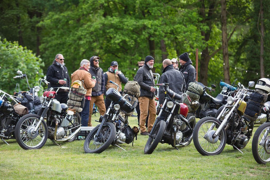 Eine Gruppe von Personen in Jacken und Helmen steht und unterhält sich in der Nähe von mehreren geparkten Motorrädern auf einer Wiese mit Bäumen im Hintergrund. An einigen Motorrädern sind Taschen oder Ausrüstung festgeschnallt.
