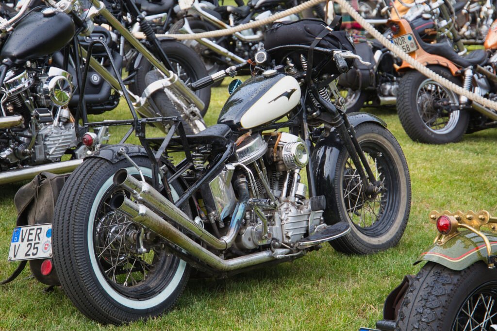 Ein schwarz-weißes Oldtimer-Motorrad mit verchromten Details parkt auf dem Rasen zwischen anderen klassischen Motorrädern bei einem Treffen oder einer Ausstellung im Freien.