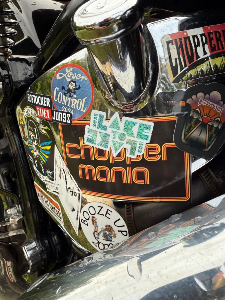 Eine Nahaufnahme eines Motorrads, das mit verschiedenen bunten Aufklebern bedeckt ist, darunter solche mit der Aufschrift "CHOPPER MANIA", "BOOZE UP SUCKERS" und "LAKE 2 LAKE". Die verchromten und schwarzen Teile des Motorrads sind sichtbar.