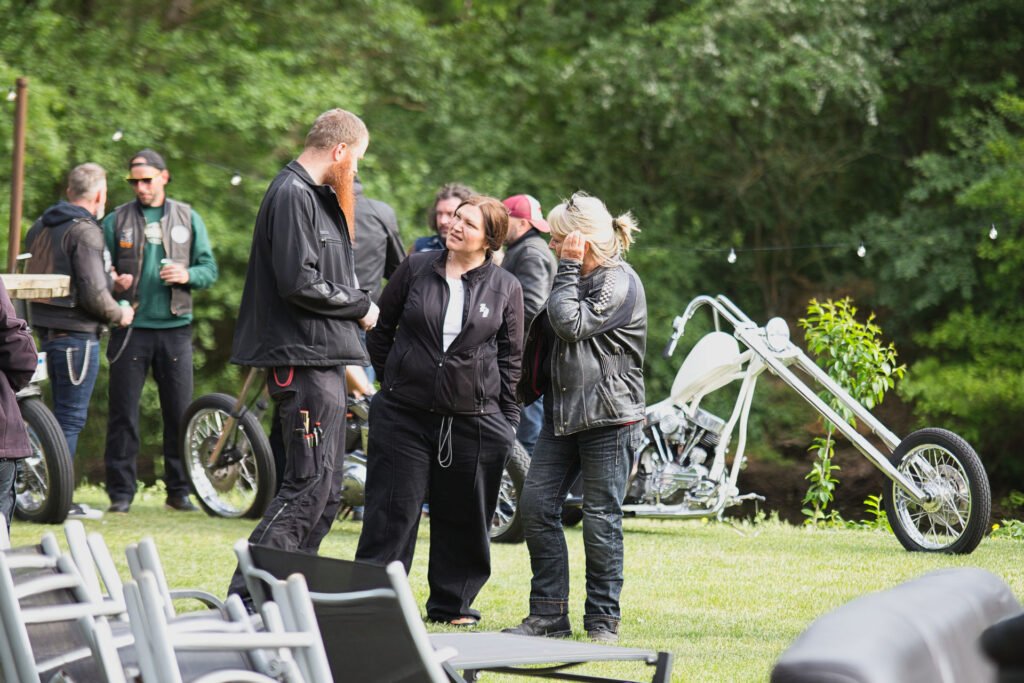 Eine Gruppe von Menschen in Freizeit- und Lederkleidung steht auf einer Wiese in der Nähe von Custom-Motorrädern und unterhält sich, während im Hintergrund Bäume und Gartenstühle zu sehen sind.