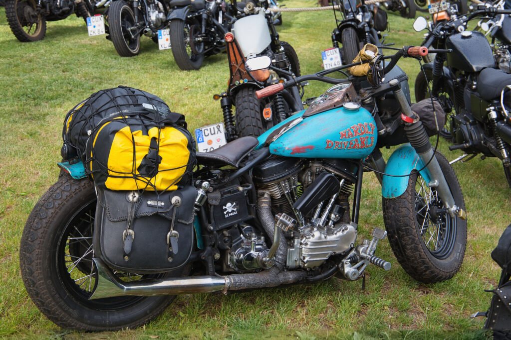 Ein türkisfarbenes Harley-Davidson-Motorrad mit Satteltaschen, einschließlich eines gelben Rucksacks, steht auf einer Wiese zwischen anderen Motorrädern bei einem Treffen.