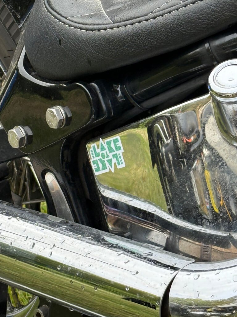 Nahaufnahme eines Motorradmotors mit einem schwarzen Sitz darüber und glänzenden Chromteilen. Auf dem Motor ist ein grüner Aufkleber mit der Aufschrift "LAKE LOVE" angebracht. Auf dem verchromten Auspuffrohr sind Wassertropfen zu sehen.