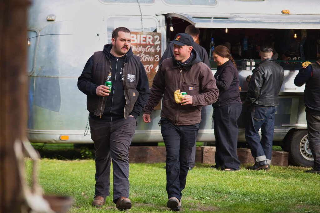 Zwei Männer gehen spazieren und unterhalten sich, während sie Getränke und Essen vor einem glänzenden Imbisswagen in der Hand halten, während andere Leute am Fenster bestellen. Die Szene findet im Freien auf einer Wiese statt.