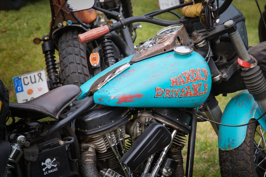 Nahaufnahme eines blauen Motorrads mit abgenutztem Lack und der Aufschrift "HARDLY DRIVABLE" in Rot und Gelb auf dem Benzintank, das auf einer Wiese zwischen anderen Motorrädern geparkt ist.