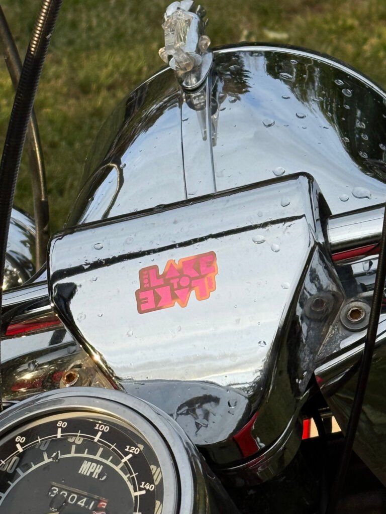 Nahaufnahme eines chromglänzenden Motorrad-Armaturenbretts mit Regentropfen auf der Oberfläche, einem Tachometer und einem rosa-gelben Aufkleber mit der Aufschrift "LAKE TO LAKE", der auf dem Kopf steht. Im Hintergrund ist Gras zu sehen.
