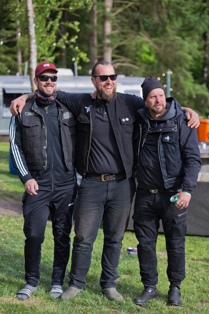 Drei Männer in schwarzen Jacken und mit Sonnenbrillen stehen im Freien, legen ihre Arme umeinander, lächeln und posieren für ein Foto auf einer Wiese mit Bäumen und Campingausrüstung im Hintergrund.
