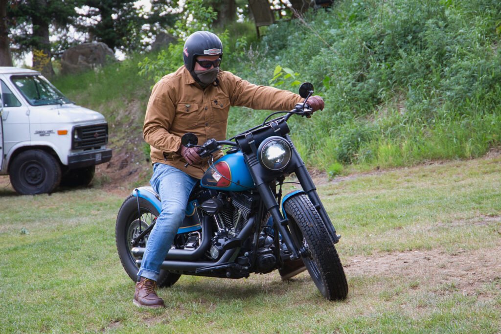 Eine Person, die einen Helm, eine Sonnenbrille, eine braune Jacke und Jeans trägt, fährt auf einem blauen Motorrad auf einer Wiese, mit einem weißen Lieferwagen und grünen Bäumen im Hintergrund.