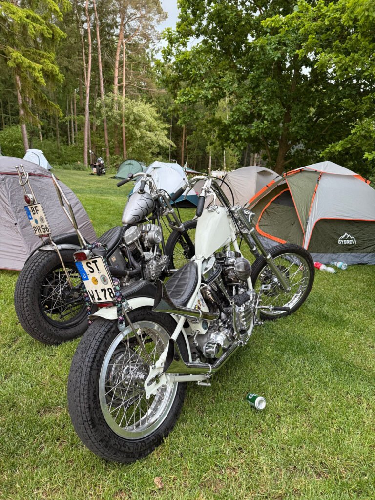 Zwei Custom-Motorräder mit deutschen Nummernschildern sind auf dem Rasen neben grauen und orangefarbenen Zelten auf einem bewaldeten Campingplatz abgestellt. Leere Dosen und Flaschen liegen in der Nähe auf dem Boden verstreut. Im Hintergrund sind Bäume und Grünflächen zu sehen.