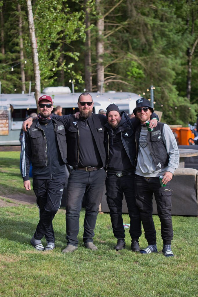 Vier Männer in schwarzen Jacken, Hosen und Sonnenbrillen stehen zusammen im Freien auf einer Wiese und legen lächelnd die Arme umeinander. Im Hintergrund sind Bäume und ein silberner Wohnwagen zu sehen.