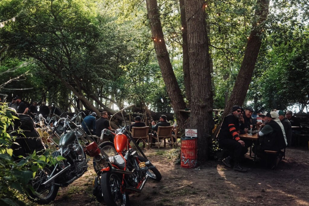 Mehrere Motorräder sind zwischen Bäumen geparkt, während Gruppen von Menschen an Tischen im Freien sitzen und sich in einem schattigen Waldstück unterhalten. Das Sonnenlicht dringt durch die Blätter und schafft eine entspannte, lockere Atmosphäre.