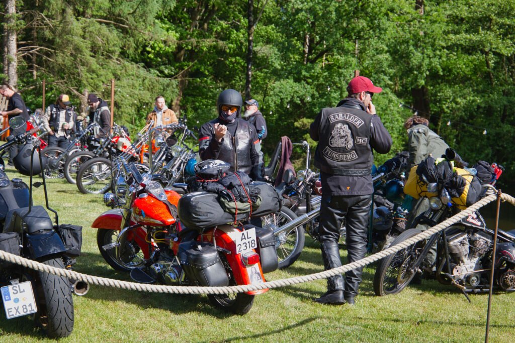 Eine Gruppe von Motorradfahrern versammelt sich im Freien zwischen geparkten Motorrädern auf einer von Bäumen umgebenen Wiese. Eine Person trägt eine rote Mütze und eine Bikerjacke, andere tragen Helme und Lederkleidung.