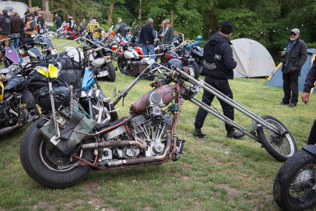 Ein Custom-Chopper-Motorrad mit einer extrem langen Vordergabel ist auf dem Rasen eines Bikertreffens im Freien geparkt, umgeben von Menschen, anderen Motorrädern und Zelten im Hintergrund.