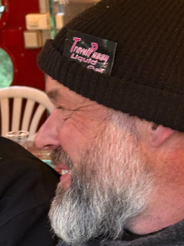 Ein bärtiger Mann, der eine schwarze Strickmütze mit einem Aufnäher trägt, auf dem "Travel Pussy Liquid Gel" steht, lächelt und blickt zur Seite. Hinter ihm sind weiße Plastikstühle und ein roter Hintergrund zu sehen.