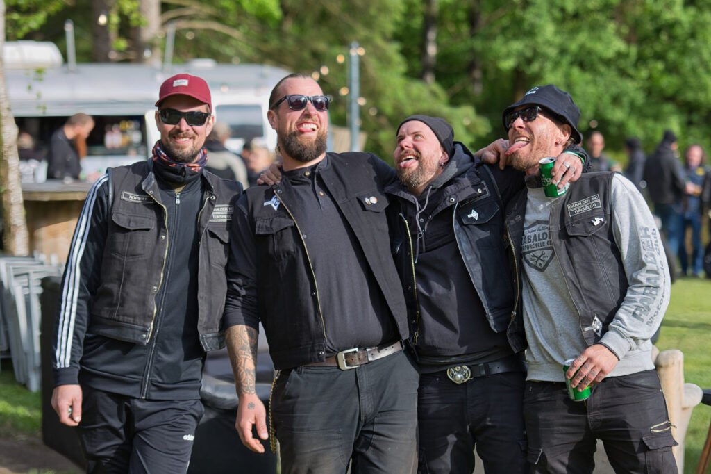 Vier Männer mit schwarzen Westen und Sonnenbrillen stehen im Freien, lächeln und lachen zusammen. Ein Mann hält ein Getränk in der Hand. Sie haben ihre Arme umeinander gelegt und scheinen ein lebhaftes, gesellschaftliches Ereignis in einer parkähnlichen Umgebung zu genießen.