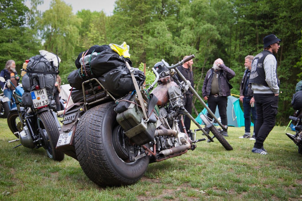 Mehrere Custom-Motorräder mit Gepäck sind auf einer Wiese geparkt, umgeben von Menschen in Freizeit- und Bikerkleidung, die sich unterhalten und in der Nähe von Bäumen und Zelten auf einer Outdoor-Veranstaltung oder einem Camping-Event stehen.