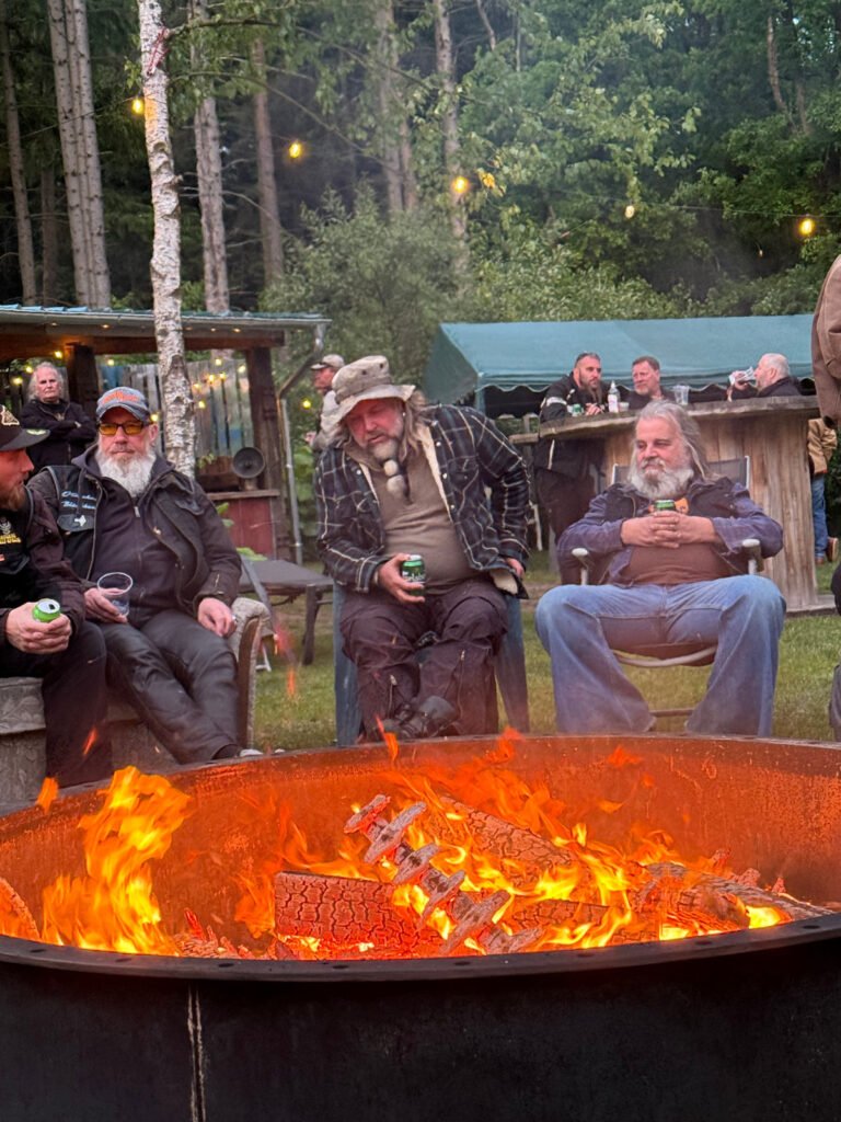 Drei ältere Männer mit Bärten sitzen um eine große Feuerstelle im Freien und halten Getränke in der Hand. Sie tragen legere, robuste Kleidung. Im Hintergrund sind Menschen und rustikale Strukturen in einer bewaldeten Umgebung mit Lichterketten zu sehen.