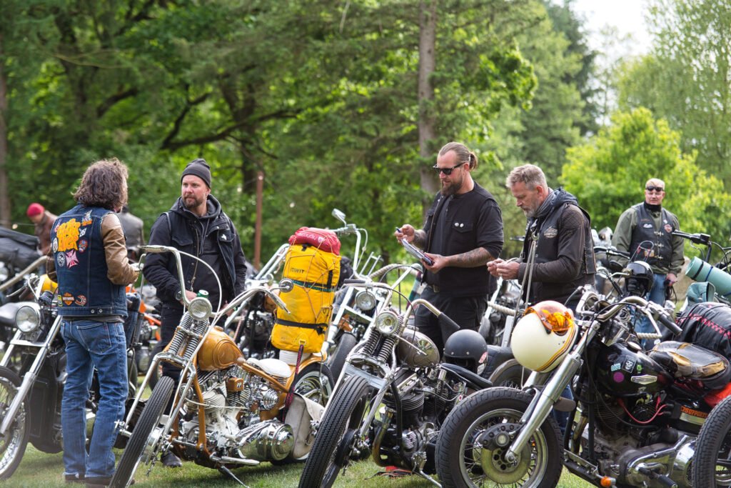 Eine Gruppe von Menschen mit Bärten und Tätowierungen steht in der Nähe von speziell angefertigten Motorrädern, die auf einer Wiese geparkt und von Bäumen umgeben sind. Einige unterhalten sich, andere überprüfen ihre Telefone. Die Szene ist lässig und im Freien.