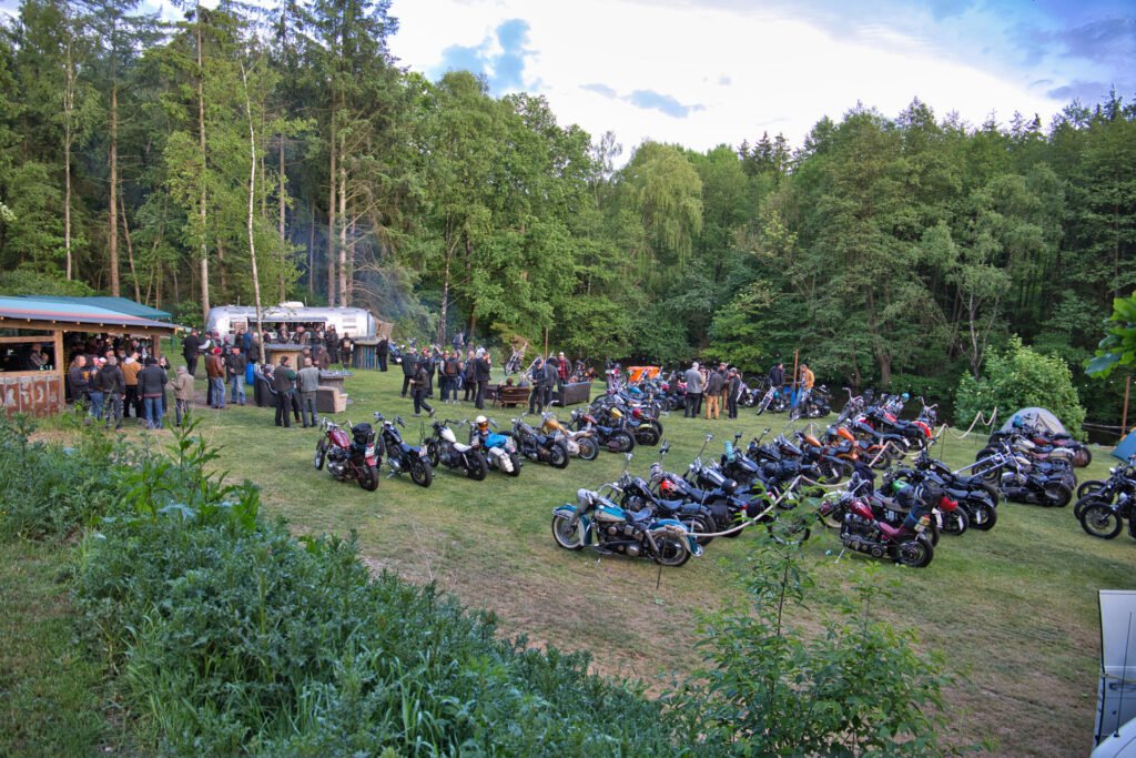 Eine Gruppe von Menschen versammelt sich im Freien in der Nähe eines Waldes, mit Reihen von geparkten Motorrädern auf einer Wiese. Einige Leute stehen in der Nähe eines kleinen Gebäudes und von Imbisswagen und unterhalten sich in einer entspannten, waldigen Umgebung.