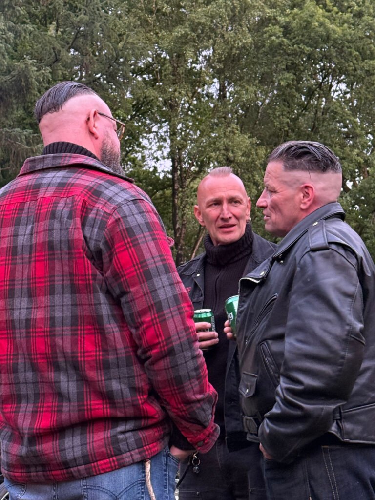 Drei Männer mit kurzen, geglätteten Frisuren stehen im Freien und unterhalten sich. Zwei von ihnen tragen schwarze Lederjacken, während der dritte eine rotkarierte Jacke trägt. Jeder von ihnen hält ein Dosengetränk in der Hand, im Hintergrund sind Bäume zu sehen.