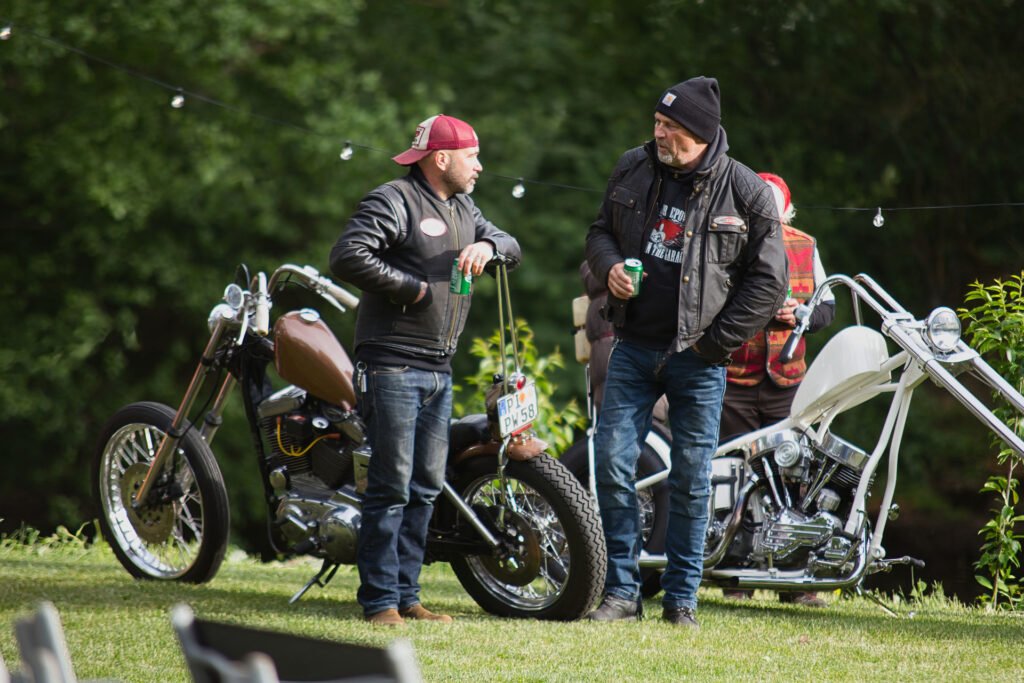 Zwei Männer in Lederjacken und Mützen unterhalten sich auf einer Wiese in der Nähe von zwei Custom-Motorrädern, wobei ein Mann ein Getränk in der Hand hält. Im Hintergrund sind Bäume und Lichterketten zu sehen.