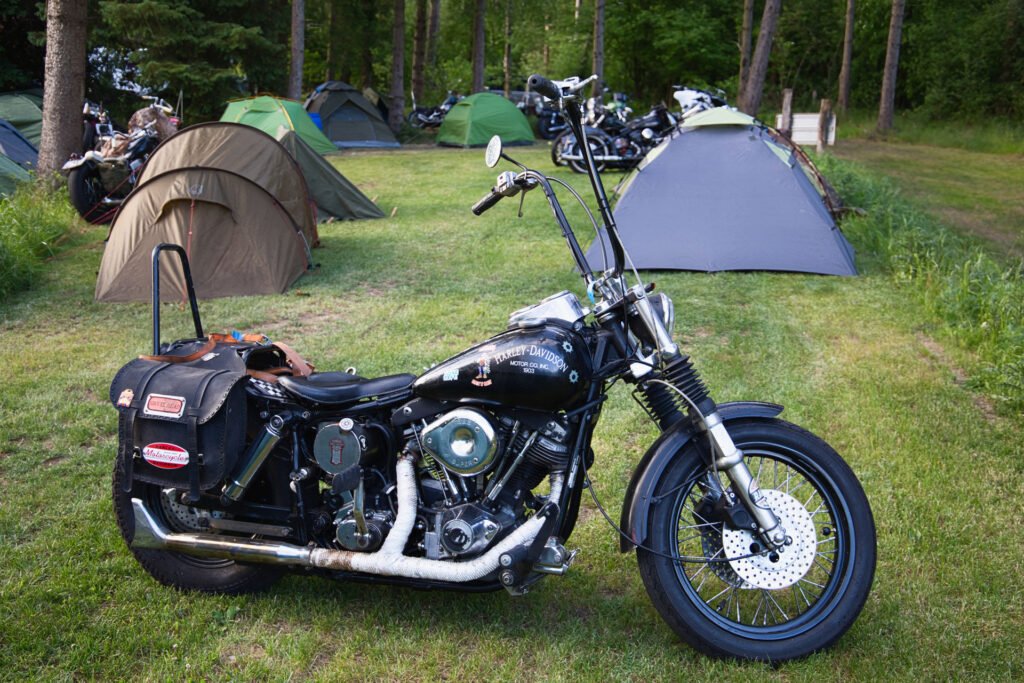 Ein schwarzes Motorrad mit Satteltaschen steht auf einer Wiese neben mehreren aufgestellten Zelten auf einem bewaldeten Campingplatz, im Hintergrund sind weitere Motorräder und Bäume zu sehen.