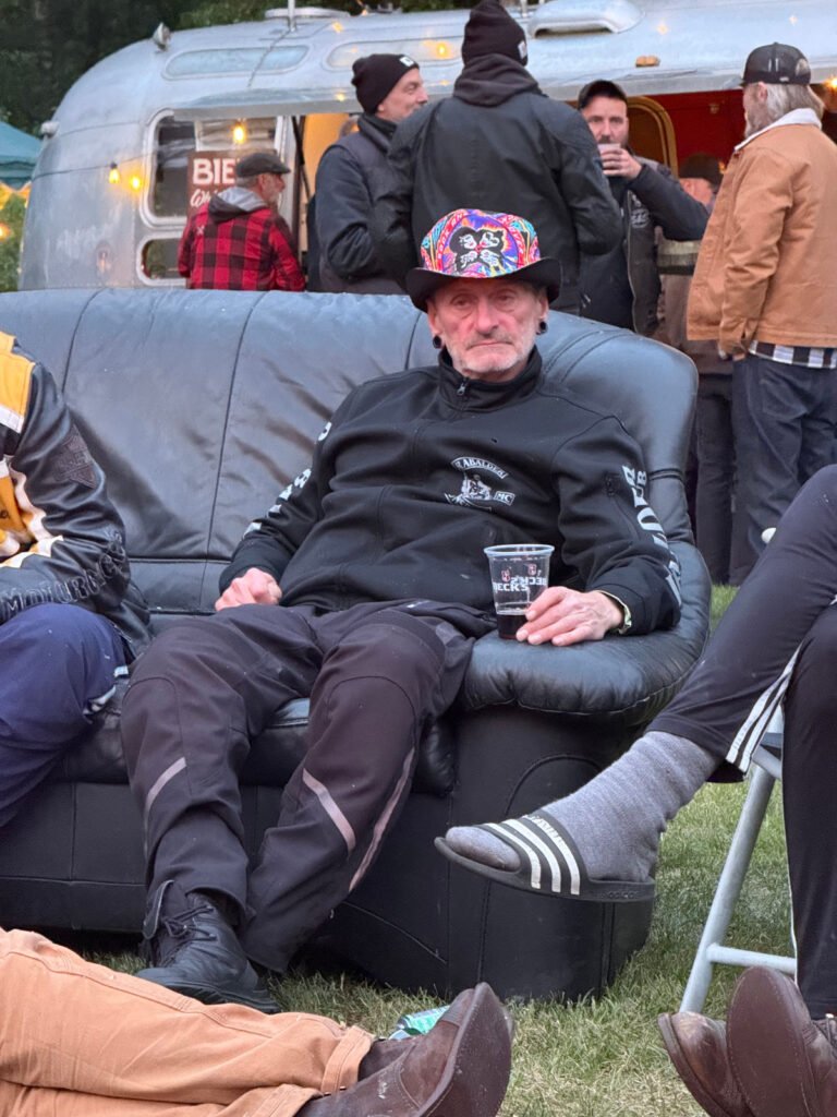 Ein älterer Mann mit einem bunten Hut sitzt auf einem schwarzen Sofa im Freien und hält ein Getränk in der Hand. Andere Leute stehen und sitzen um ihn herum, und im Hintergrund ist ein silberner Wohnwagen zu sehen. Die Szene scheint bei einer zwanglosen Zusammenkunft im Freien zu spielen.