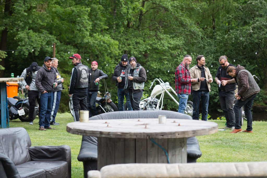 Eine Gruppe von Männern, von denen einige Lederjacken und Bikerwesten tragen, steht im Freien in der Nähe von Motorrädern auf einer Wiese zusammen, mit Bäumen im Hintergrund und Gartenmöbeln im Vordergrund.