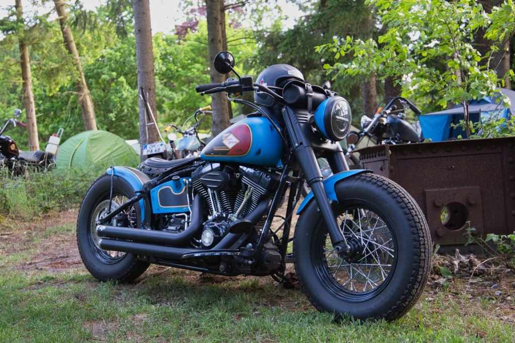Ein blau-schwarzes Motorrad im Vintage-Stil ist auf einer Wiese in der Nähe von Bäumen und einem grünen Zelt geparkt, während im Hintergrund eine Campingausrüstung und ein weiteres Motorrad zu sehen sind.
