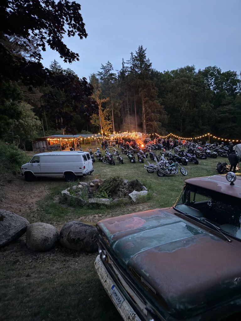 Eine Gruppe von Menschen versammelt sich in der Abenddämmerung um ein Feuer auf einer grasbewachsenen Lichtung mit geparkten Motorrädern, Lichterketten, einem weißen Lieferwagen und einem rostigen grünen Lastwagen, umgeben von Bäumen.