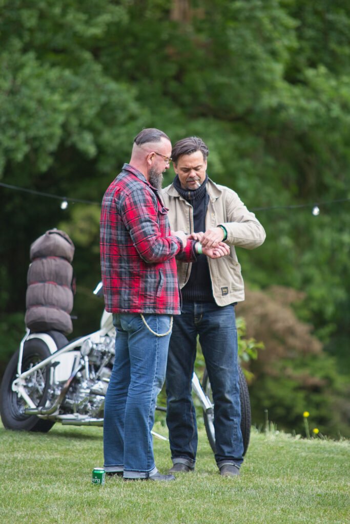 Zwei Männer stehen im Gras, der eine in einem rotkarierten Hemd, der andere in einer beigen Jacke. Sie schauen gemeinsam auf eine Uhr. Ein Motorrad und eine Dose sind in der Nähe, mit grünen Bäumen im Hintergrund.
