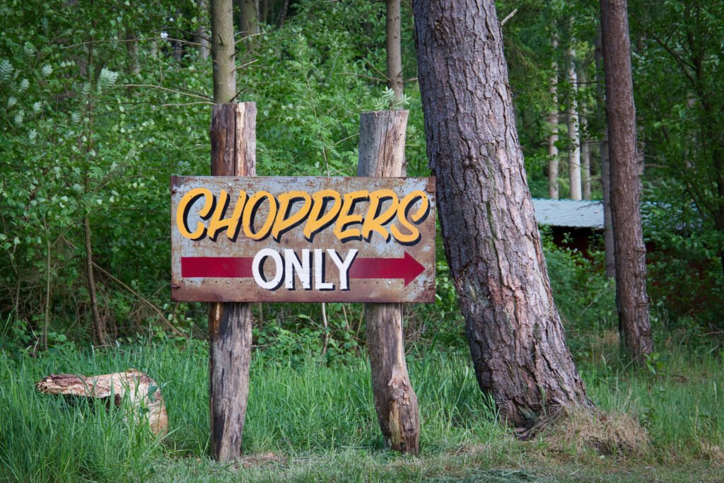 Auf einem Holzschild in einem Waldgebiet steht "CHOPPERS ONLY" mit einem Pfeil, der nach rechts zeigt. Hohe Bäume und grünes Gras umgeben das Schild.