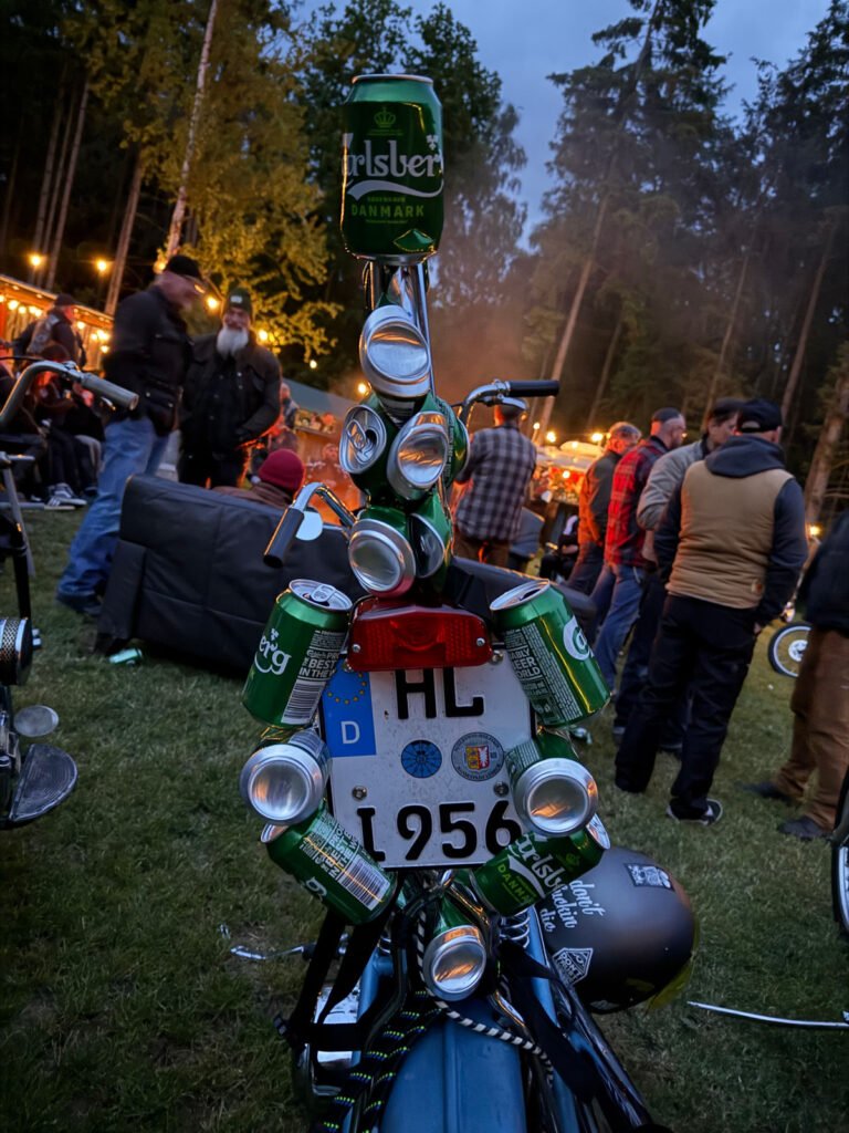 Das Heck eines Motorrads ist mit zahlreichen leeren Carlsberg-Bierdosen verziert, die über dem Nummernschild angeordnet sind. Die Szene spielt im Freien bei einem nächtlichen Treffen mit Menschen und Lichterketten im Hintergrund.