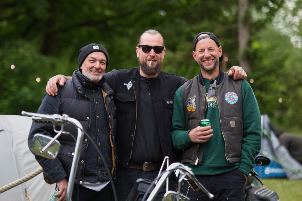 Drei Männer in legerer Kleidung im Biker-Stil stehen im Freien, legen die Arme umeinander und lächeln in die Kamera. Im Vordergrund sind zwei Motorräder zu sehen, und im Hintergrund stehen Bäume.