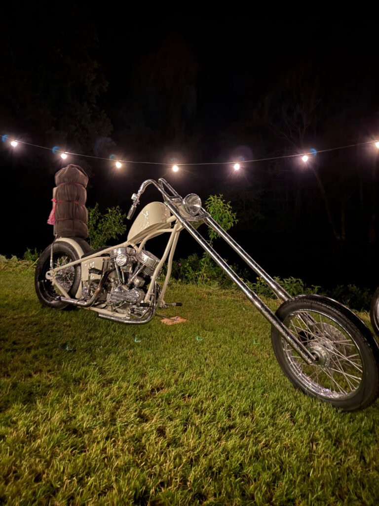 Ein Custom-Chopper-Motorrad mit verlängerter Vordergabel ist nachts auf einer Wiese geparkt und wird von einer Lichterkette über dem Kopf beleuchtet. Bäume und Dunkelheit füllen den Hintergrund.