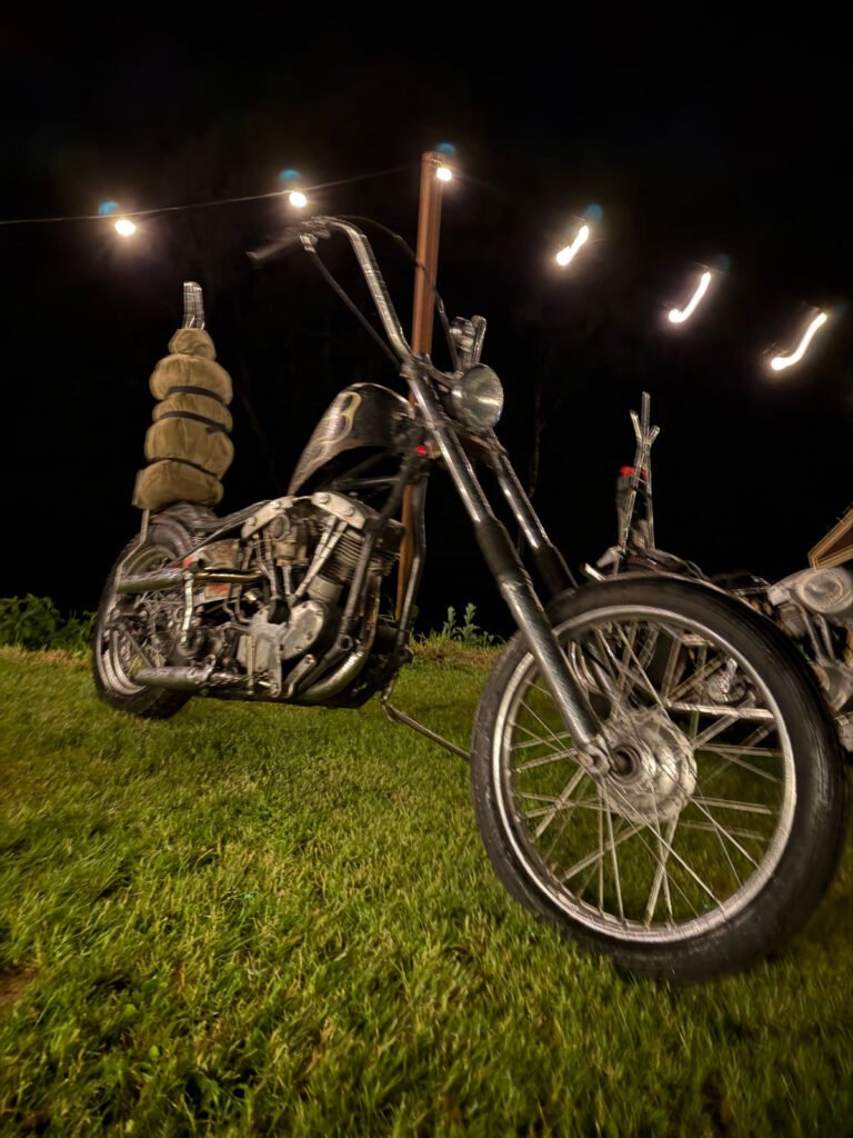 Ein schwarzes Motorrad mit hohem Lenker ist nachts auf einer Wiese unter Lichterketten geparkt. Ein anderes Motorrad ist teilweise im Hintergrund zu sehen. Die Szene ist im Freien und leicht unscharf.