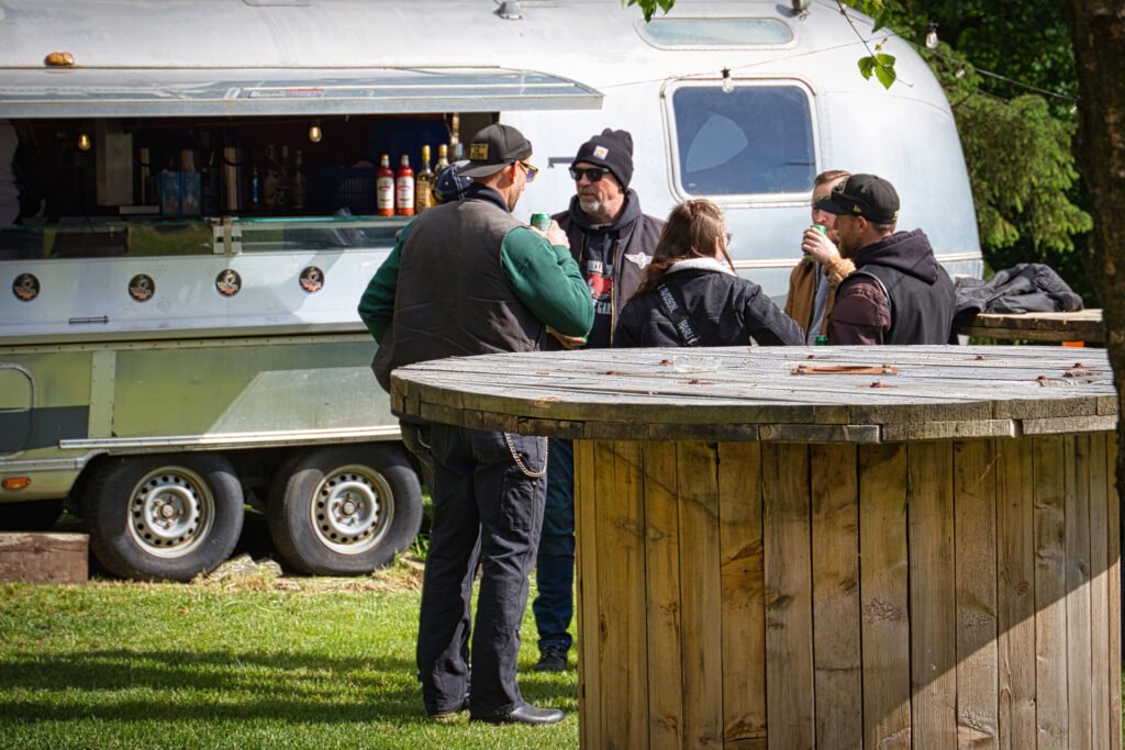 Vier Personen stehen und unterhalten sich an einem großen hölzernen Spulentisch im Freien, während im Hintergrund ein silberner Anhänger als Essens- oder Getränkestand dient. Die Gruppe ist mit Jacken und Hüten bekleidet.