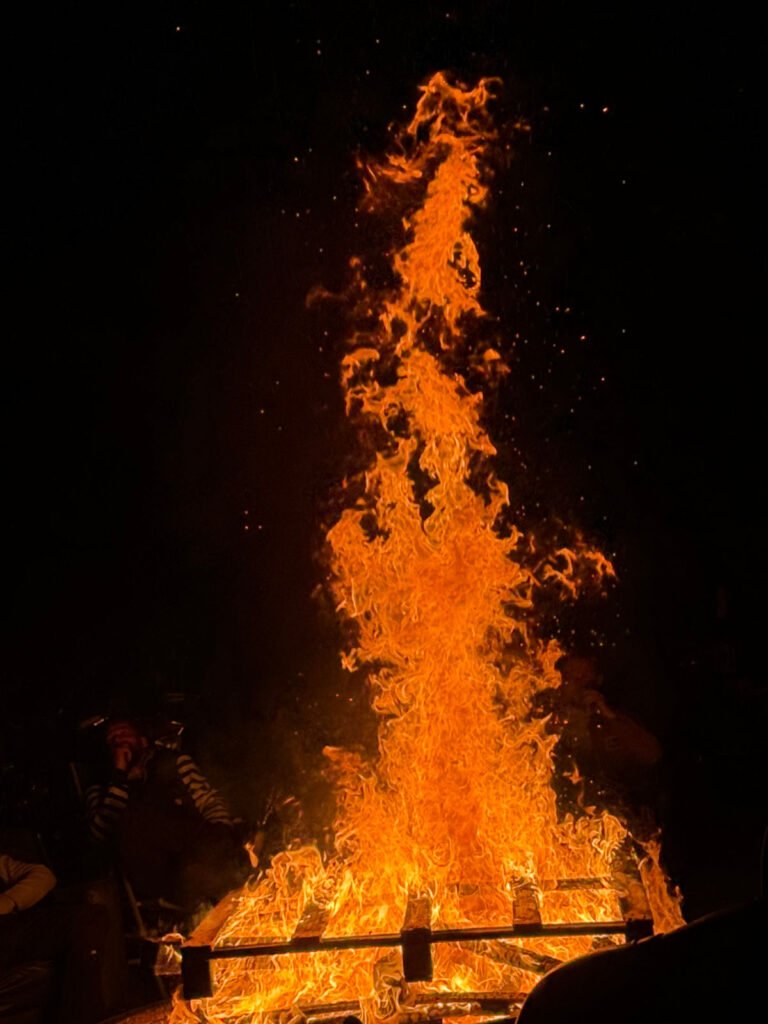 Ein großes, leuchtend orangefarbenes Lagerfeuer lodert vor einem dunklen Hintergrund in die Höhe, und um das Feuer herum sitzen Menschen, die teilweise im Schatten sichtbar sind.